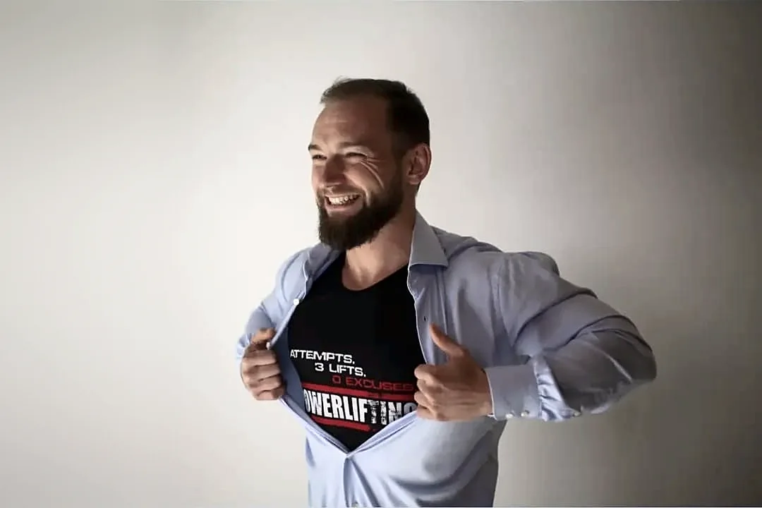 Uśmiechnięty Marcin Bielicki rozchyla koszulę, pod którą ma koszulkę z napisem Powerlifting